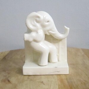 Pode incluir: Estatueta de elefante em cerâmica cor marfim, sentada e encostada em um suporte retangular. O elefante tem detalhes, incluindo uma tromba, orelhas e pernas. A estatueta é um item decorativo.