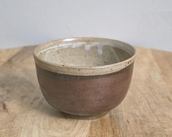 Op de afbeelding: Een kleine keramische kom met een tweekleurig ontwerp, voorzien van een mat donkerbruin onderste gedeelte en een gespikkelde crèmekleurige rand. De binnenzijde van de kom is eveneens crèmekleurig gespikkeld met een ronde vorm en een voet.