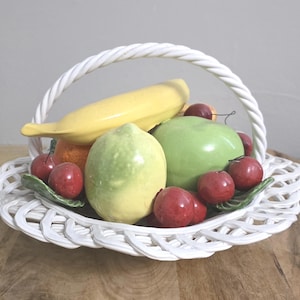 Op de afbeelding: Een witte keramische mand met een gevlochten latwerkontwerp met een verzameling kunstfruit, waaronder een gele banaan, een lichtgroene citroen, een gladde groene appel en verschillende kleine, glanzende rode kersen met groene blaadjes.