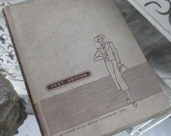 Op de afbeelding: Een vintage schoolboek met een beige linnen kaft. De kaft toont een lijntekening van een persoon in een pak en hoed, met een handtas. De tekst "TEXT EDITION" staat in een bruin ovaal en de McGraw-Hill Book Company Inc. is onderaan gedrukt.