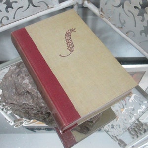 Puede incluir: Un libro antiguo de tapa dura con una cubierta de lino beige y un lomo rojo. La cubierta presenta un diseño de espiga de trigo marrón.