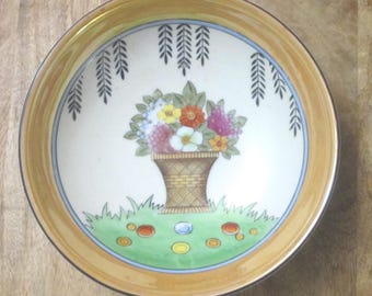 Op de afbeelding: Een ronde keramische kom met een brede, goudbruine rand en een centrale illustratie van een gevlochten mand vol kleurrijke bloemen. Het ontwerp bevat groen gras, kleine ronde accenten en hangende zwarte wilgentakken.
