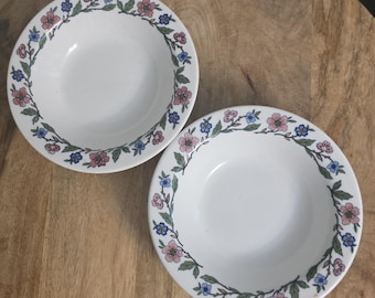 Op de afbeelding: Twee witte keramische kommen met een doorlopend randpatroon van roze en blauwe bloemen met groene bladeren. De kommen hebben een gladde, ronde binnenkant en een brede, platte rand versierd met een delicaat bloemmotief rondom het midden.