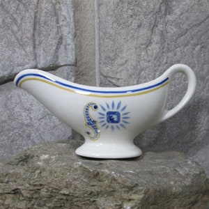 May include: White ceramic gravy boat with a blue and gold design featuring a stylised seahorse and a sunburst. The boat has a handle on the right side.