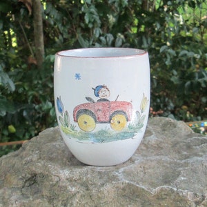 May include: A white ceramic mug with a hand-painted design of a boy driving a red car. The car has yellow wheels and the boy is wearing a blue hat. The mug has a brown rim.