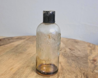 Op de afbeelding: Een doorzichtige glazen fles met een zwarte dop, voorzien van reliëffiguren op het bovenste gedeelte. Het etiket vermeldt Mémoire chérie, Flower Mist, Elizabeth Arden en 4 fl. oz. De bodem bevat een kleine hoeveelheid amberkleurige vloeistof.