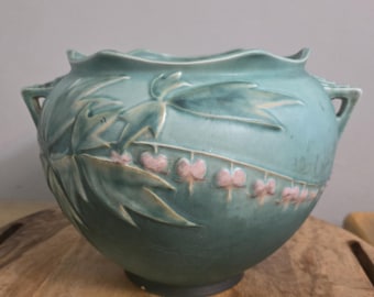 Op de afbeelding: Een matte, blauwgroene keramische kom met een geschulpte rand en twee handvatten aan de zijkanten. De buitenkant heeft een reliëfontwerp van groene bladeren en een horizontale rij kleine, lichtroze hartvormige bloemen rond het midden.