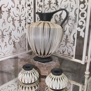 May include: Three black and white woven ceramic vases with a decorative design. The vases are on a white surface with a white metal screen in the background.