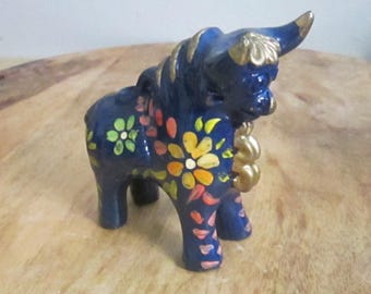 Könnte beinhalten: Dunkelblaue Keramik-Stierfigur, verziert mit bunten Blumenmustern in Gelb, Orange und Rosa. Hörner und Mähne des Stiers sind goldfarben akzentuiert. Ein dekoratives Stück.