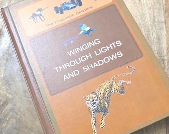 Op de afbeelding: Een bruin gebonden boek getiteld Winging Through Lights and Shadows uit de serie The Linguistic Readers. De kaft toont een springende jachtluipaard, een klein figuurtje met een blauwe wagen, een raceauto, een hond en een persoon in een jas.