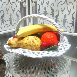 May include: White ceramic fruit basket with a yellow banana, a pear, an orange, and red cherries. The basket is decorated with a white woven pattern.