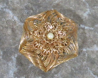 Könnte beinhalten: Eine goldfarbene Brosche mit einem filigranen, kunstvollen Blumendesign. In der Mitte der Blüte befindet sich eine kleine, weiße Perle.