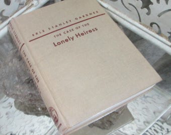 Könnte beinhalten: Ein beigefarbenes Taschenbuch mit dem Titel "The Case of the Lonely Heiress" von Erle Stanley Gardner. Das Buch liegt auf einer weißen Oberfläche mit einem dekorativen Metalldesign.