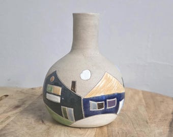 Könnte beinhalten: Eine runde Keramikvase mit engem Hals, die eine handbemalte Szene mit zwei Häusern in Schwarz-, Blau- und Gelbtönen zeigt. Die Grundfarbe ist ein mattes Beige, und das Design umfasst kleine quadratische Fenster sowie eine weiße Wolke.