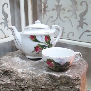 Op de afbeelding: Een wit porseleinen theeservies met een bloemenmotief. De theepot en het theekopje hebben roze rozen en groene bladeren.