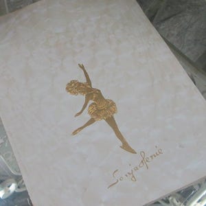 May include: A white card with a gold foil image of a ballerina in a tutu. The card is signed "Sonja Henie".