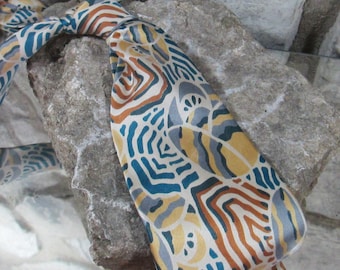 Op de afbeelding: Een patroon stropdas met een knoop, met een ontwerp van turquoise, bruine, gele en grijze vormen. De stropdas is gedrapeerd over een ruw, grijs stenen oppervlak, met het ingewikkelde patroon en de kleuren.