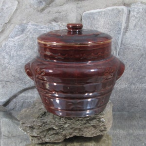 Può includere: Un vaso di ceramica marrone con un coperchio, che presenta un design decorativo di bordi a festoni e motivi floreali. Il vaso ha due maniglie sui lati.