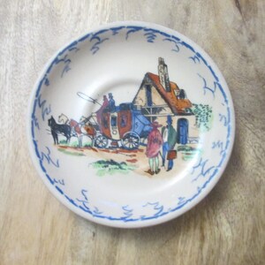 May include: A small, round, white ceramic plate with a blue scalloped border. The plate features a colorful illustration of a horse-drawn carriage, a building, and figures in period clothing. The scene is set against a light background.