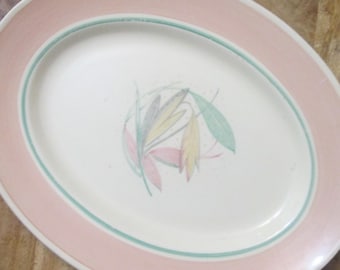 Op de afbeelding: Ovale serveerschaal met een roze rand en een groene accentlijn. Het midden van de schaal is wit met een bloemmotief in roze, geel en groen. Een vintage stuk om eten te serveren.