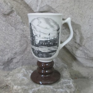 May include: A white ceramic mug with a black and white street scene illustration. The mug has a brown pedestal base and handle. The mug is designed to hold hot beverages.