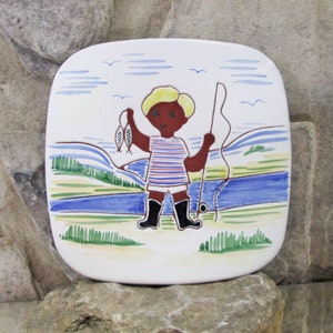 Puede incluir: Un plato cuadrado de cerámica blanca con un diseño pintado a mano de un niño pescando. El niño lleva una camisa a rayas rojas y azules, botas negras y sostiene una caña de pescar y dos peces. El fondo es un río azul con hierba verde y un cielo azul con nubes blancas.