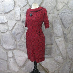 May include: A red dress with a black geometric pattern and a black collar. The dress has a button on the front and pockets on the sides.