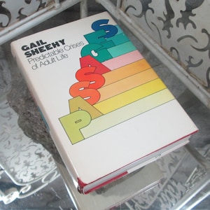 Może przedstawiać: Książka w twardej oprawie zatytułowana "Passages: Predictable Crises of Adult Life" autorstwa Gail Sheehy. Okładka zawiera tytuł dużymi, kolorowymi literami i wzór w paski w kolorach tęczy. Książka spoczywa na odblaskowej powierzchni.