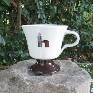 May include: White ceramic coffee cup with a brown base. The cup has a stylised letter "h" with a flame on top.