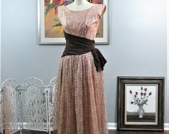 Op de afbeelding: Een vintage roze kanten jurk met een bruine fluwelen band en strik. De jurk heeft een ronde hals en korte mouwen. De jurk is op een mannequin en wordt getoond tegen een witte muur.