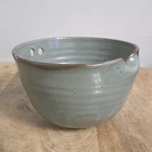 Puede incluir: Un cuenco mezclador de cerámica color verde salvia con acabado moteado y silueta redondeada. El cuenco presenta un pequeño pico vertedor en el borde y dos orificios circulares cerca del borde superior, con un borde marrón oscuro acentuado.