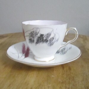 May include: A white teacup and saucer set with a gold handle and trim. The cup and saucer feature a black and burgundy floral design. The teacup is sitting on a wooden surface.