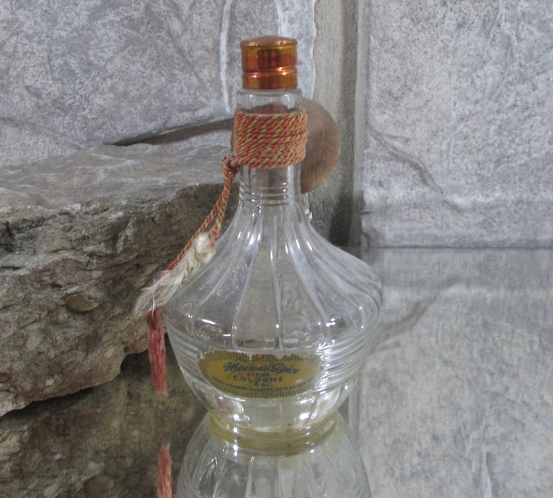 Puede incluir: Una botella de vidrio transparente con un borla roja y dorada y una etiqueta que dice "Marcella's Cologne".