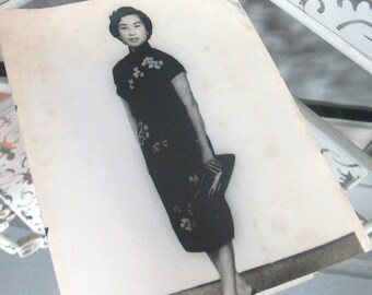 Könnte beinhalten: Schwarzweißfoto einer Frau in einem schwarzen Kleid mit Blumenstickerei. Sie hält eine kleine Handtasche in ihrer rechten Hand.
