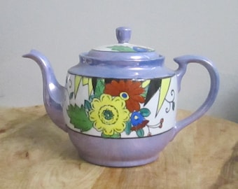 Op de afbeelding: Een vintage keramische theepot in tinten lavendel en wit, versierd met een bloemenontwerp. De theepot heeft een gebogen tuit, een handvat en een deksel met een kleine knop. Het bloemmotief omvat gele, rode en blauwe bloemen met groene bladeren.