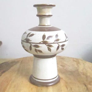 May include: A ceramic vase with a cream-colored base and a wide, rounded body featuring a brown painted leaf vine pattern. The vase has a flared neck and a solid brown circular base, with additional brown horizontal bands accenting the design.