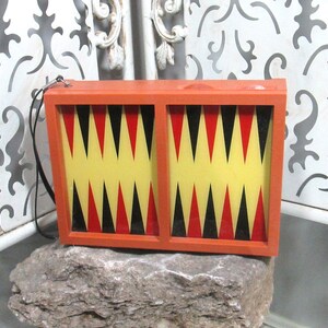 Puede incluir: Un juego de backgammon vintage de madera con un marco rojo y un tablero de damas de color amarillo, negro y rojo. El juego es de tamaño compacto y portátil y tiene un asa de transporte.