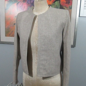 Puede incluir: Una chaqueta corta de color gris claro con manga larga y cierre de un botón. La chaqueta está hecha de una tela texturizada.