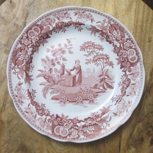 May include: A white ceramic plate with a scalloped edge and a detailed maroon floral border. The center features a pastoral scene with a figure near a well. The plate is likely vintage or antique, suitable for display or dining.