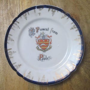 Può includere: Piatto in ceramica bianca con bordo blu e dettagli dorati. Il piatto presenta uno stemma con le parole "Present from Poole" e un bordo decorativo. Il piatto è su una superficie di legno.