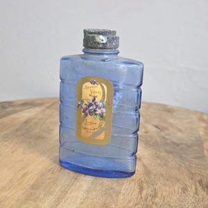May include: A rectangular blue glass bottle with horizontal ridges and a weathered grey cap. A gold-toned label on the front features a graphic of purple violet flowers and the text Special Violet, Lynas, Logansport, Ind.