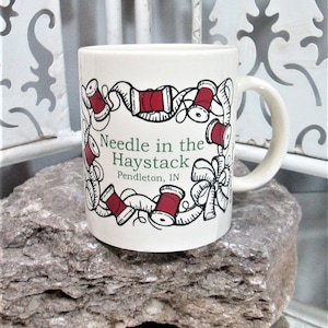 Może przedstawiać: Biały kubek ceramiczny z czarno-czerwonym wzorem szpulki nici i miarki. Na kubku nadrukowany jest tekst "Needle in the Haystack Pendleton, IN".