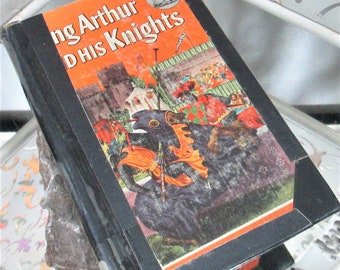 Op de afbeelding: Een vintage hardcover boek getiteld "King Arthur and His Knights" met een oranje omslag en een illustratie van ridders te paard. Het boek heeft een zwarte rug en een zwarte rand rond de omslag.