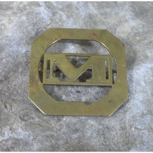 Może przedstawiać: Złota metalowa spinka z geometrycznym wzorem przedstawiającym literę "M" w środku. Spinka ma kształt kwadratu ze zaokrąglonymi rogami.