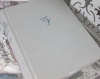 Op de afbeelding: Een lichtgrijs hardcover boek met de initialen "EG" in turquoise script op de omslag. De rug van het boek toont gedeeltelijke tekst. Het boek ligt op een oppervlak met een decoratief patroon.