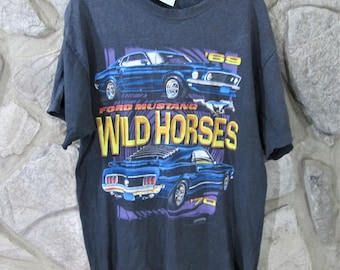 Op de afbeelding: Een zwart T-shirt met een vintage afbeelding van een blauwe Ford Mustang. De afbeelding bevat de tekst "Ford Mustang Wild Horses" en het jaar "1969".