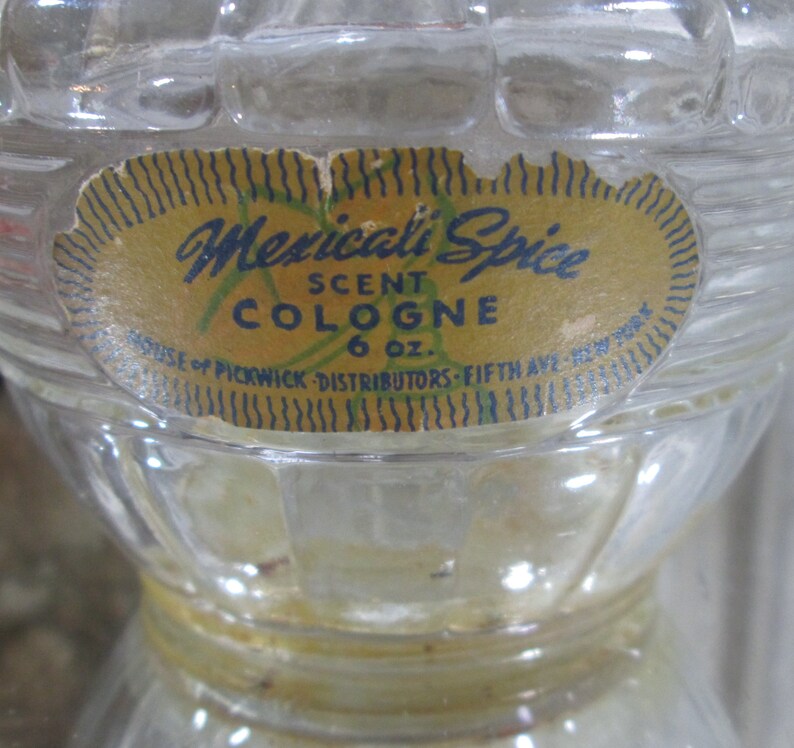 Puede incluir: Una botella de vidrio transparente con una superficie texturizada. La botella tiene una etiqueta amarilla con ribete azul y negro. La etiqueta dice "Mexicali Spice Scent Cologne 6 oz. House of Pickwick Distributors - Fifth Ave. New York."