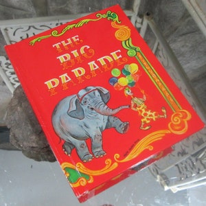 Puede incluir: Un libro de tapa dura rojo con una ilustración colorida de un elefante y un payaso. El título del libro es "The Big Parade" con las palabras "with monkeys" en una fuente más pequeña debajo del título.