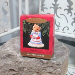 May include: A red Hallmark Keepsake box featuring a teddy bear in a blue dress and white apron holding a tray of cookies. The box has a gold oval label and the text "THE BEARINGERS". The box is on a gray rock.