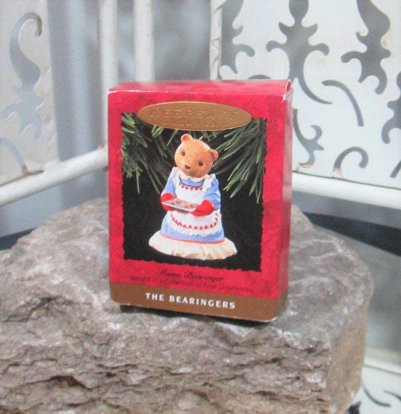 May include: A red Hallmark Keepsake box featuring a teddy bear in a blue dress and white apron holding a tray of cookies. The box has a gold oval label and the text "THE BEARINGERS". The box is on a gray rock.
